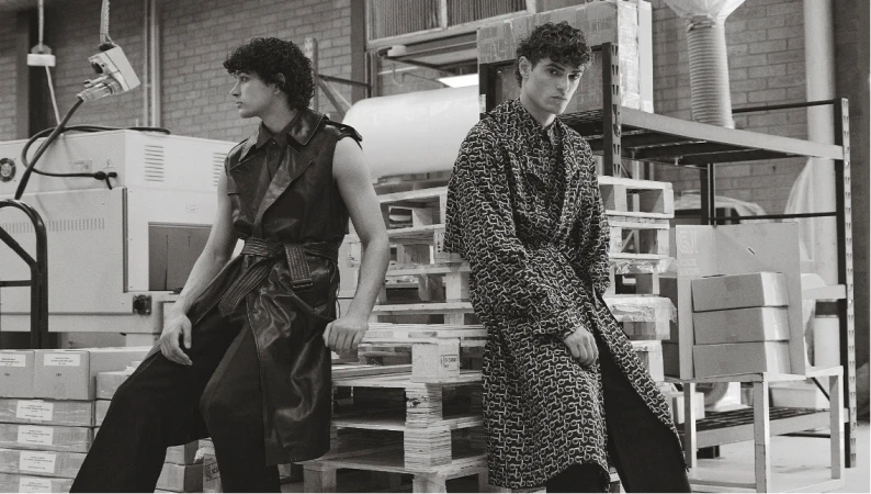 Two adult men in patterned coats, posing on a wooden pallet in an industrial setting, creating a high-fashion editorial image.