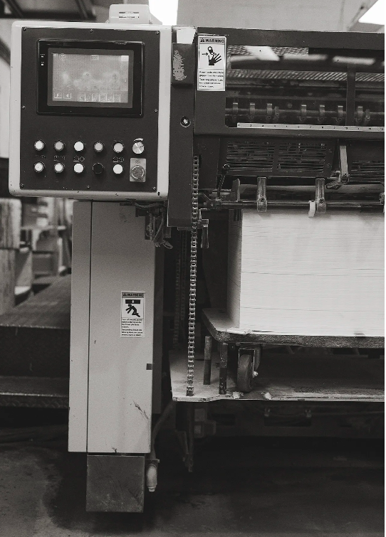 Industrial printing machinery showing the control panel, paper feed, warning labels, metal components, and various mechanical elements.