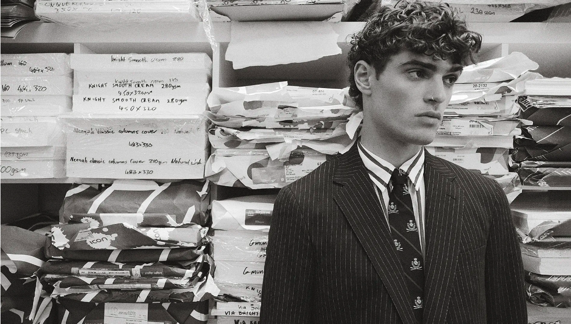 Young man in pinstripe suit and tie poses in a warehouse with packaged goods on shelves, creating a high-fashion, editorial-style photo.