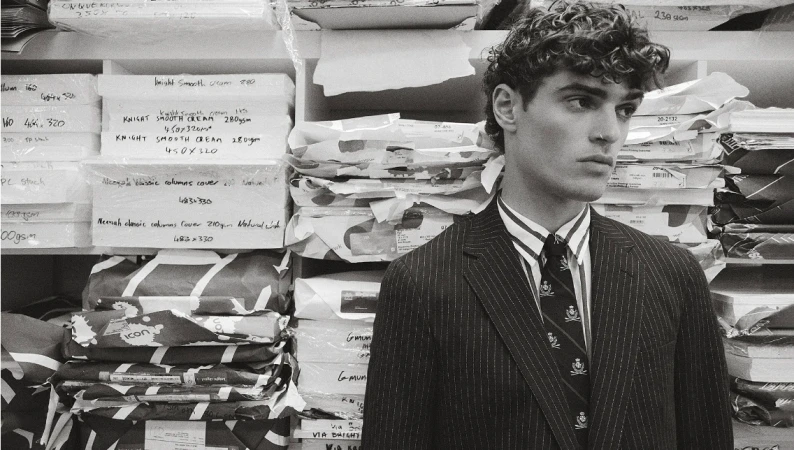 Young man in pinstripe suit and tie poses in a warehouse with packaged goods on shelves, creating a high-fashion, editorial-style photo.