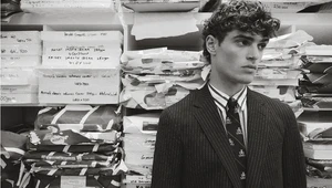 Young man with curly hair wearing a pinstripe suit and tie standing against shelves filled with paper stocks.