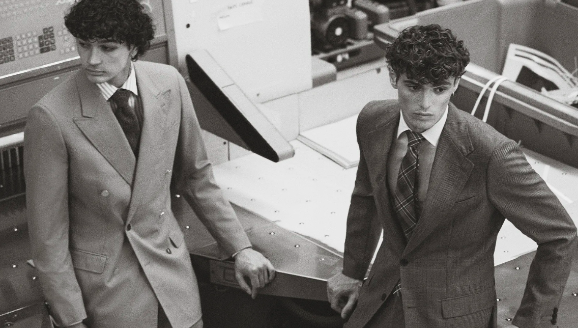 Two adult men in suits pose indoors, showcasing formal wear in a monochromatic palette with sharp tailoring and classic styling.