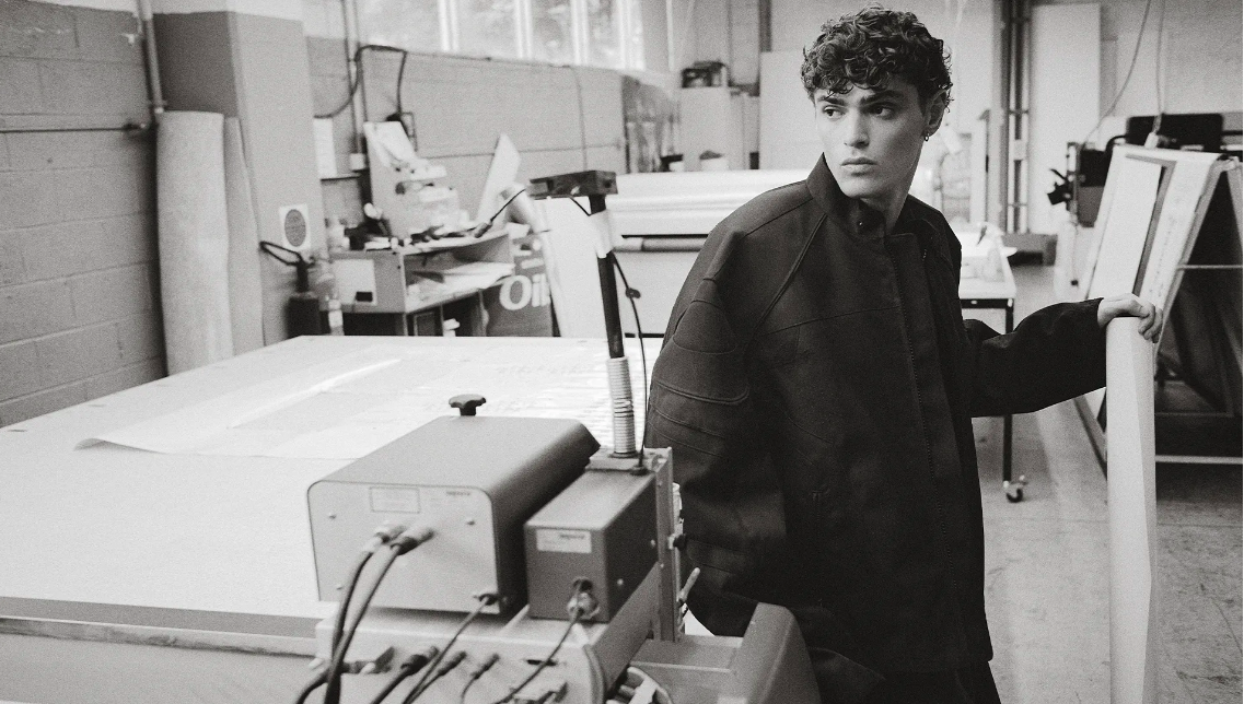 Monochrome portrait of a young man with curly hair in a jacket, standing indoors near equipment, looking away from the camera.