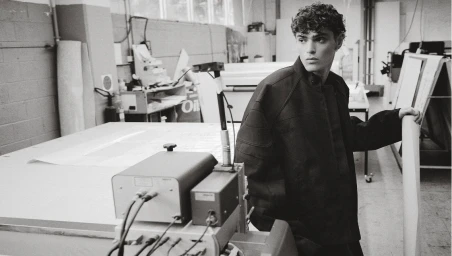 Young man in dark jacket standing in a workshop with large printing equipment for Icon Magazine Australia
