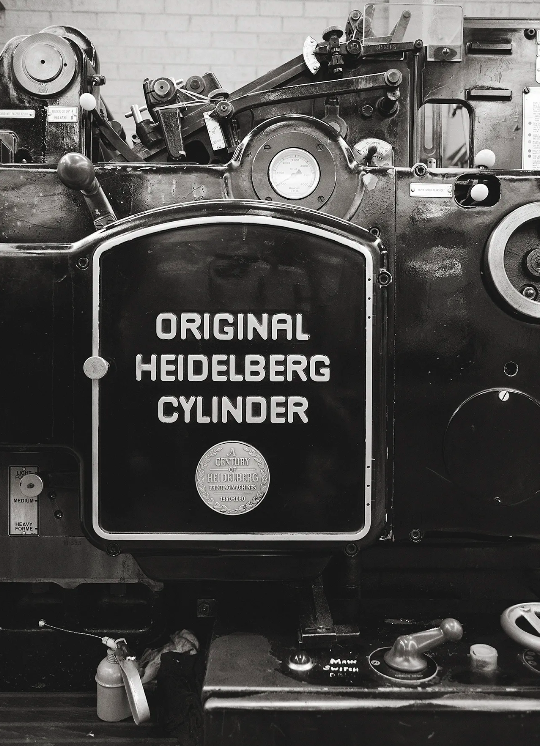 Monochrome image of a vintage Heidelberg cylinder printing press machine in a workshop, close-up view of machinery and nameplate.