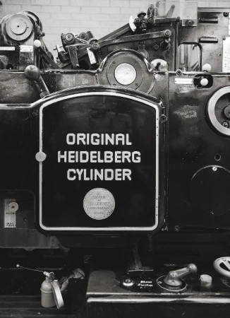Monochrome image of a vintage Heidelberg cylinder printing press machine in a workshop, close-up view of machinery and nameplate.