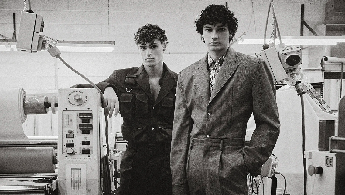 Monochrome studio shot of two adult males wearing suits next to industrial sewing machine, evoking a vintage, fashion-forward aesthetic.