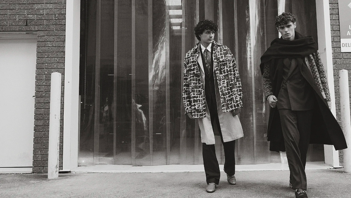 Two adult males walk side-by-side in patterned coats on an urban street, captured in a black and white fashion editorial photograph.