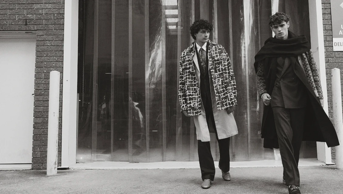 Two adult males walk side-by-side in patterned coats on an urban street, captured in a black and white fashion editorial photograph.