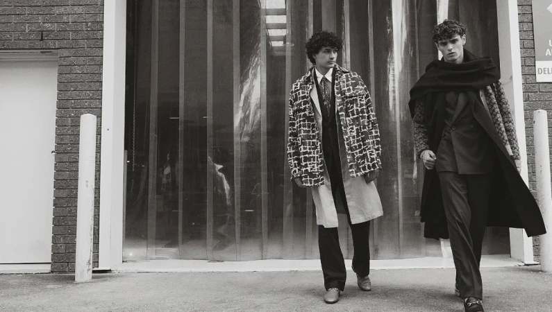 Two adult males walk side-by-side in patterned coats on an urban street, captured in a black and white fashion editorial photograph.