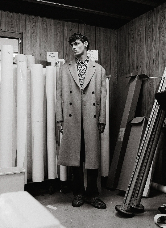Monochrome full shot of a curly-haired male model wearing a patterned shirt, double-breasted coat and loafers in an indoor studio setting.