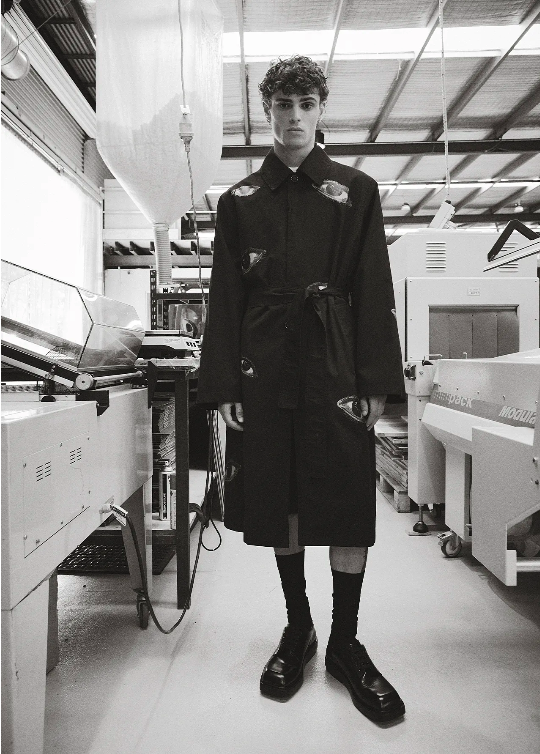 A person posing in a black coat in an industrial setting, with machinery visible in the background and wearing socks and shoes.