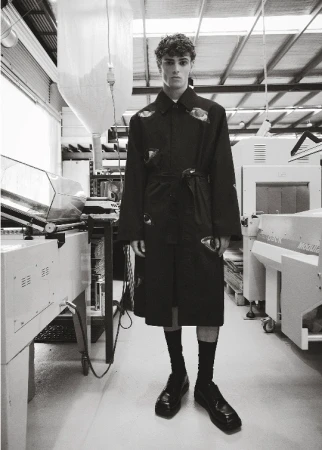 A person posing in a black coat in an industrial setting, with machinery visible in the background and wearing socks and shoes.