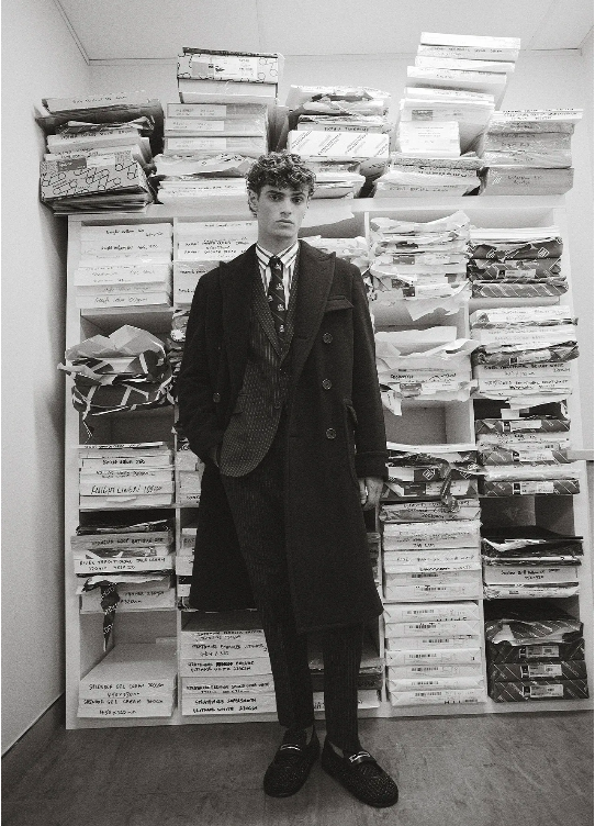 A young man in a dark coat and patterned suit stands in front of shelves filled with stacked boxes in a black and white editorial fashion shot.