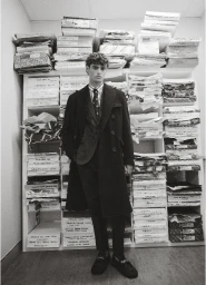 Man wearing a tailored suit and dark overcoat standing before shelves packed with stacked paper materials