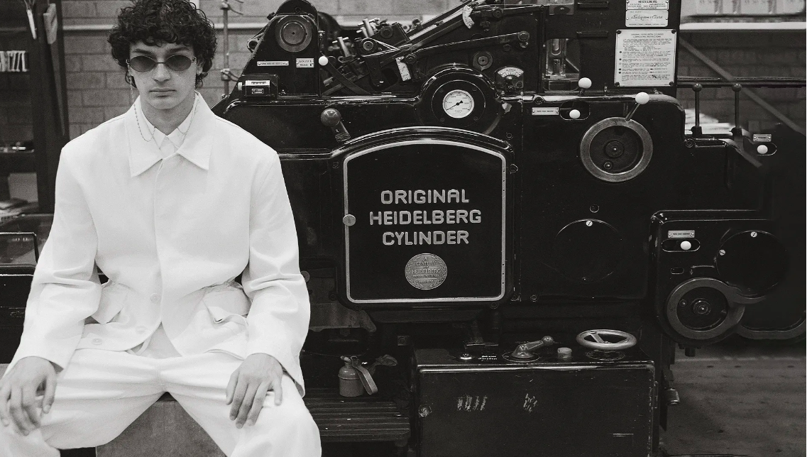 Monochrome portrait of adult male model in white suit seated in front of vintage Heidelberg Cylinder printing press machine.