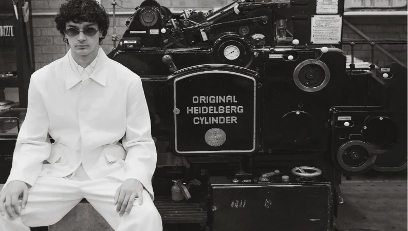 Monochrome portrait of adult male model in white suit seated in front of vintage Heidelberg Cylinder printing press machine.
