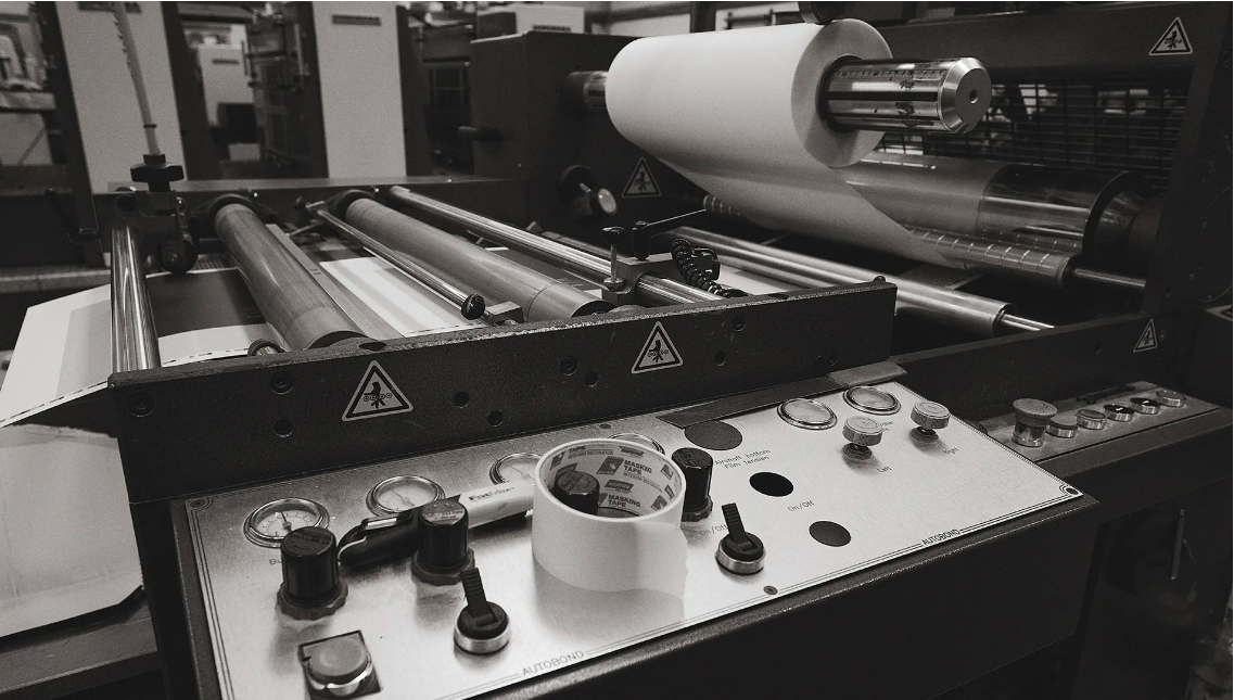 Industrial printing press with paper roll, rollers, and control panel in a monochrome setting, showcasing machinery and manufacturing process.