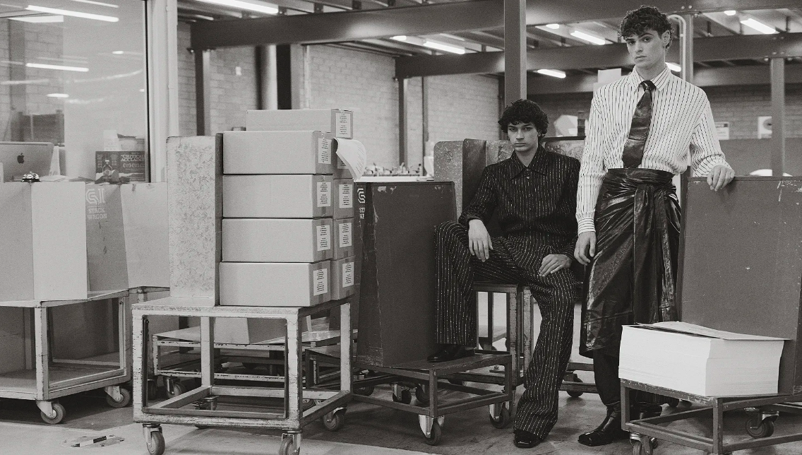 Two adult male fashion models wearing striped clothing pose in a warehouse setting with boxes and metal carts in black and white.