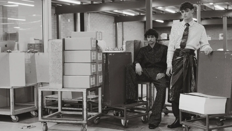 Two adult male fashion models wearing striped clothing pose in a warehouse setting with boxes and metal carts in black and white.