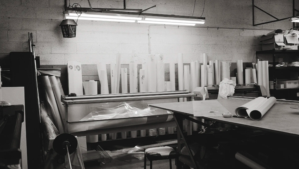 Black-and-white shot in a printing workshop featuring rolls of paper, a table, and overhead fluorescent lighting illuminating the space.