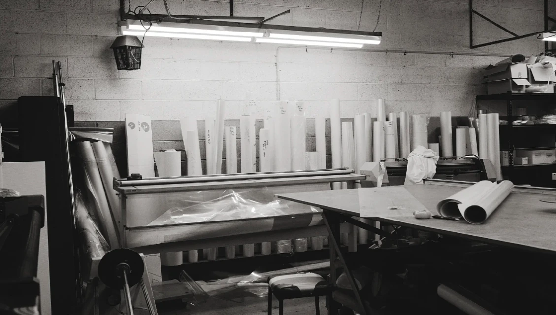 Black-and-white shot in a printing workshop featuring rolls of paper, a table, and overhead fluorescent lighting illuminating the space.