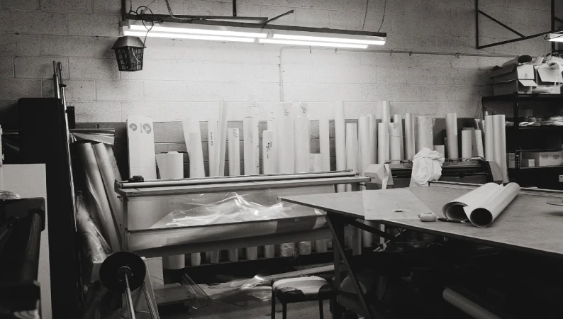Black-and-white shot in a printing workshop featuring rolls of paper, a table, and overhead fluorescent lighting illuminating the space.