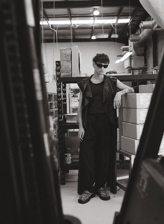 Black and white editorial photo of a young man wearing sunglasses and a leather vest in a warehouse setting