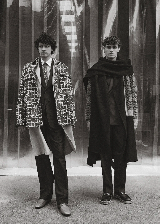 Two men in patterned overcoats and dark suits pose against a reflective panel wall in a black and white fashion shot.