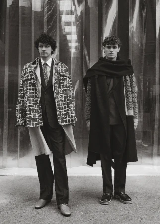 Two men in patterned overcoats and dark suits pose against a reflective panel wall in a black and white fashion shot.