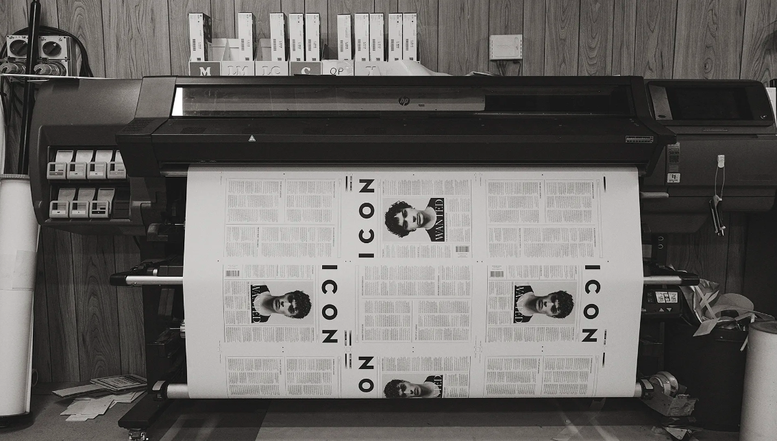 A black-and-white photograph shows a large-format printer outputting pages with 'ICON' and a man's portrait, against a wood-panel wall.
