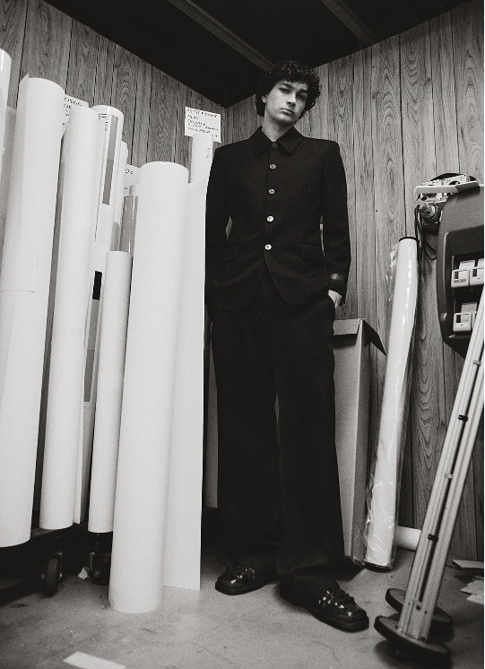 Black and white portrait of a male model in a dark suit standing in front of rolled paper in what appears to be a photo studio.