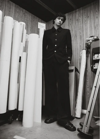 Black and white portrait of a male model in a dark suit standing in front of rolled paper in what appears to be a photo studio.