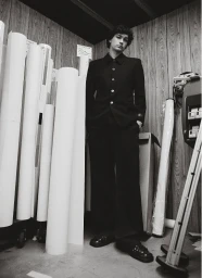 Monochrome fashion editorial image of a young man in a black suit standing in a narrow room with rolled paper tubes.