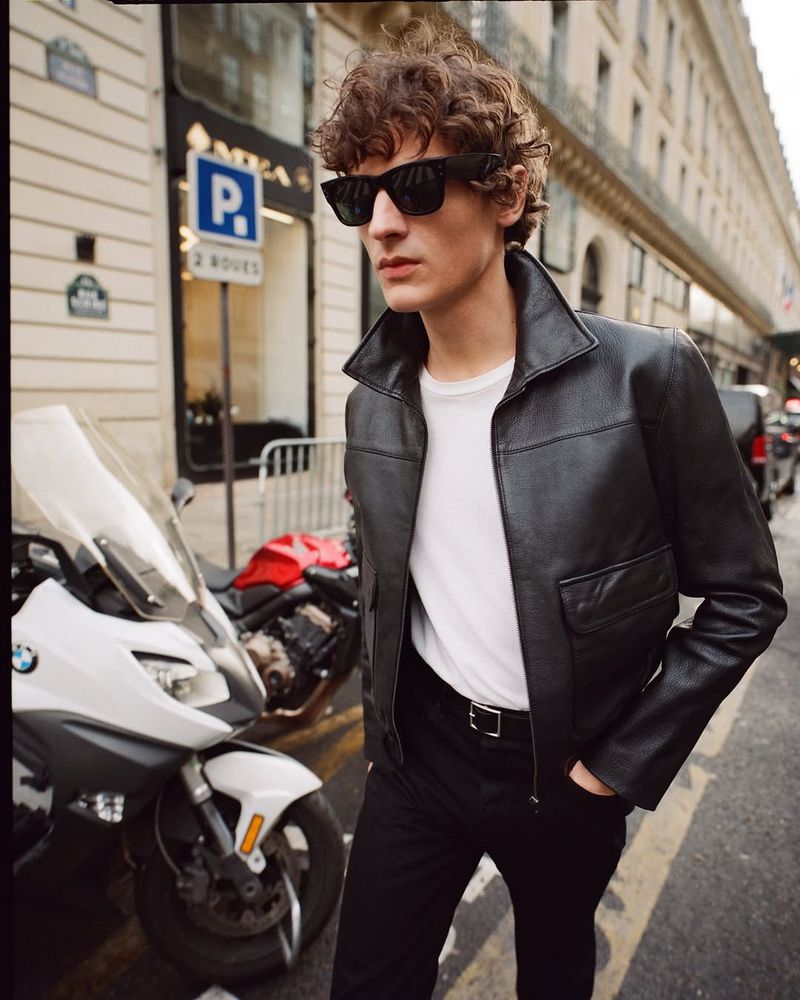 Man in black leather jacket and sunglasses walking on a city street next to parked motorcycle. Fashion editorial photography.