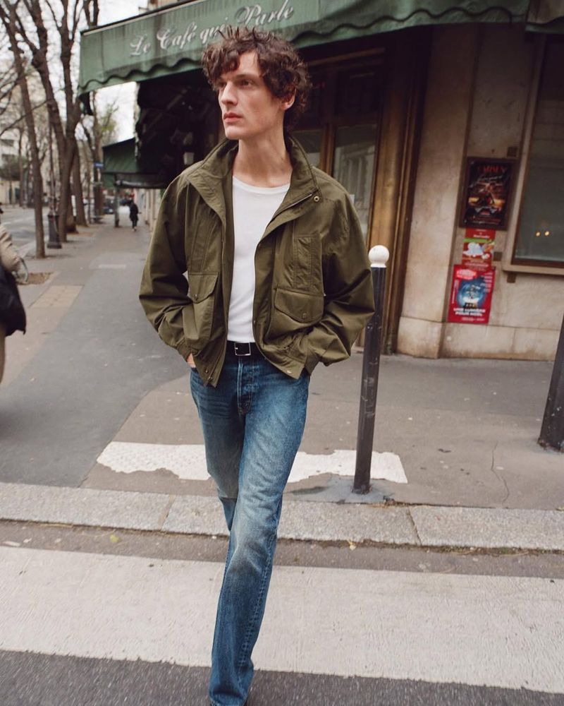Adult male with curly hair wearing a white shirt, green jacket, and jeans walking in an urban setting with storefronts and trees.