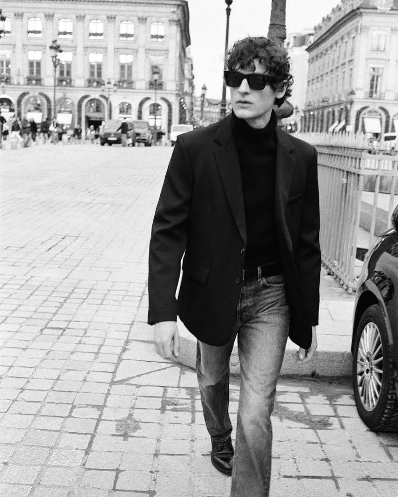 A stylish man walks on a cobblestone street in a black blazer, turtleneck, jeans, and sunglasses, in a black and white fashion photograph.
