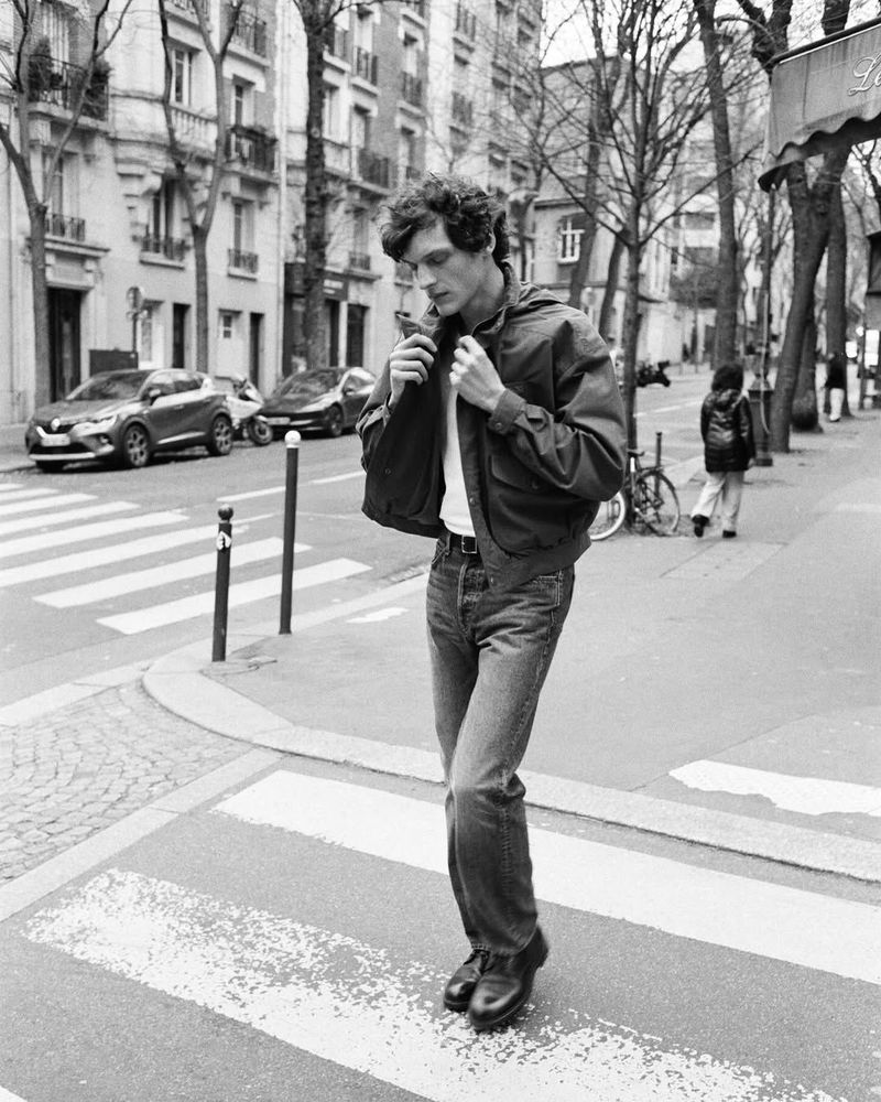 Black and white shot of a young man in jeans and jacket crossing a street in Paris, with buildings and cars in the background.