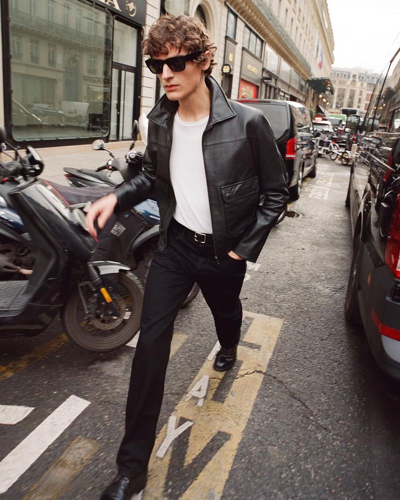 A young adult man in a black leather jacket walks on an urban street next to parked cars and a motorcycle, wearing sunglasses.