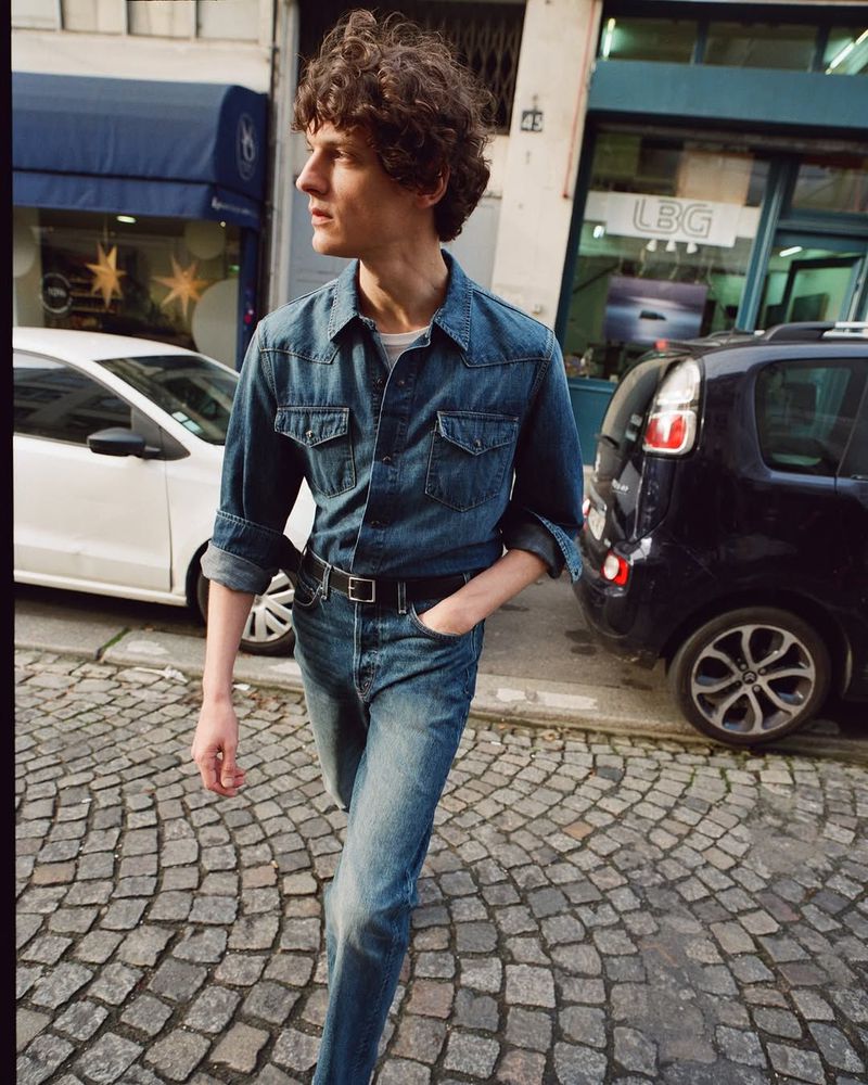 A young adult male with curly hair poses on a cobblestone street wearing a denim shirt and jeans, with cars and buildings in the background.