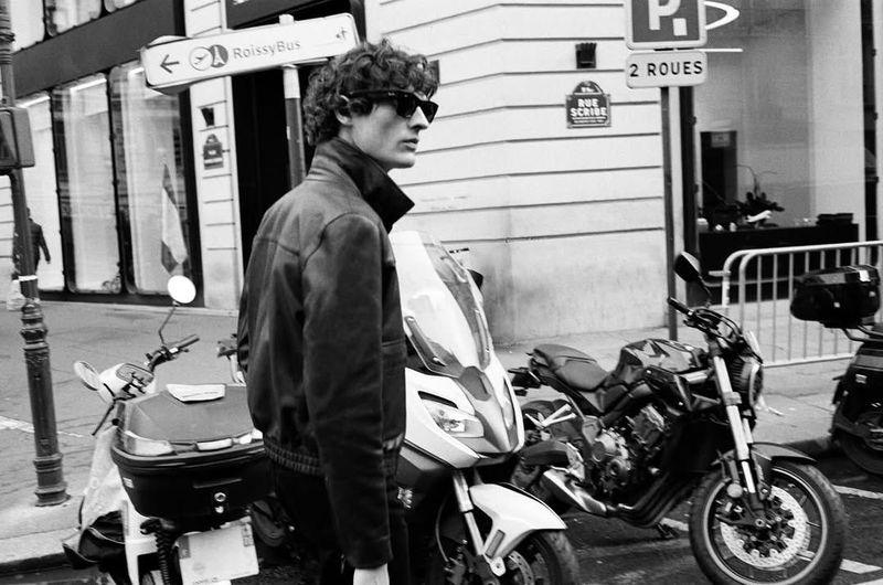 Man in leather jacket and sunglasses stands next to motorcycles on a city street with European signage in black and white.
