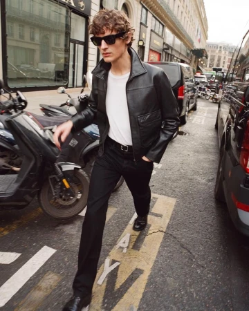 Model wearing black leather jacket, white t-shirt, and dark sunglasses walking down a crowded city street