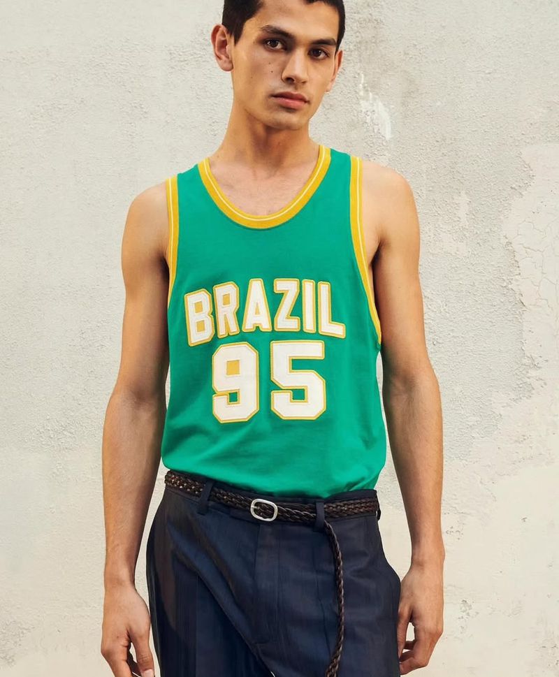 A young adult male stands in a green and yellow Brazil jersey and navy dress pants against a textured wall, in a medium indoor shot.