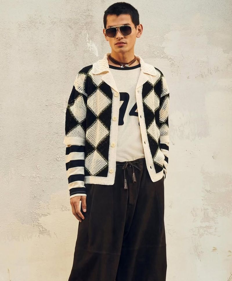 Young man in a knitted black and white cardigan with sunglasses poses against a textured wall in an eye-level medium shot.
