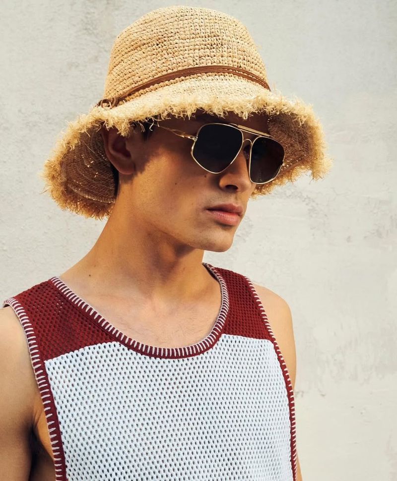 Close-up of a young adult male wearing a straw hat and sunglasses, styled in a white and burgundy tank top against a neutral background.
