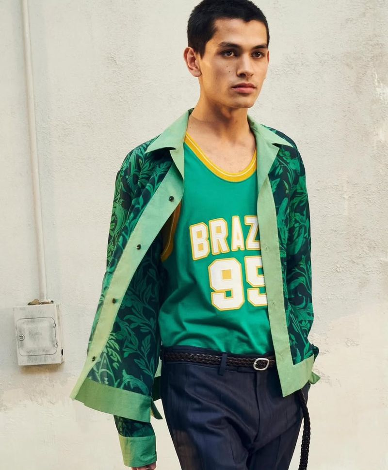 A young adult man poses in a studio wearing a green jersey and matching floral patterned jacket. He is wearing dark pants and a braided belt.