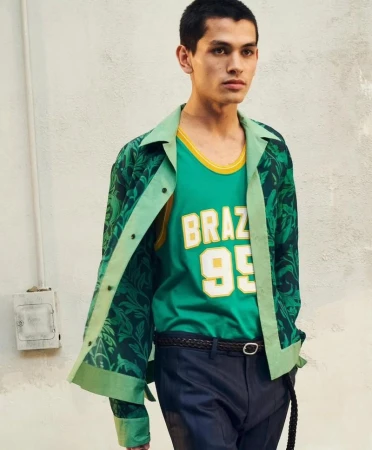 A young adult man poses in a studio wearing a green jersey and matching floral patterned jacket. He is wearing dark pants and a braided belt.