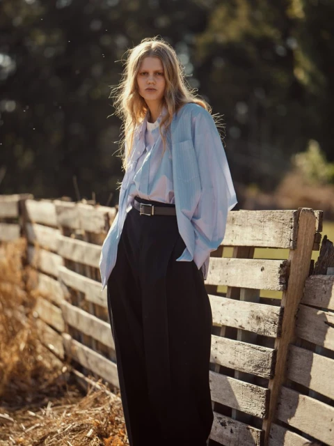 Fashionable young woman with blonde hair posing outdoors in a blue blouse, black pants, and brown belt against a wooden fence.