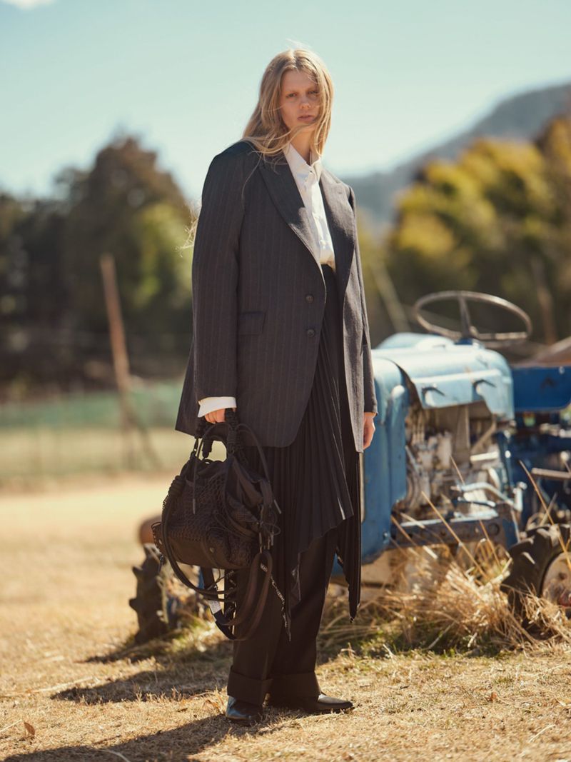 Woman in pinstripe suit with backpack stands in rural setting near blue tractor on a sunny day, fashion editorial lifestyle shot.