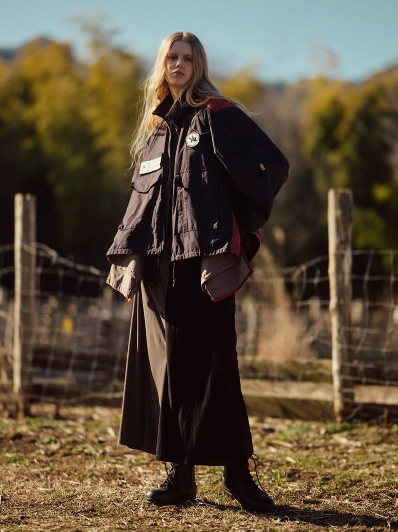 Model wearing a dark utility jacket and long skirt outdoors near a rustic wooden fence in a field.